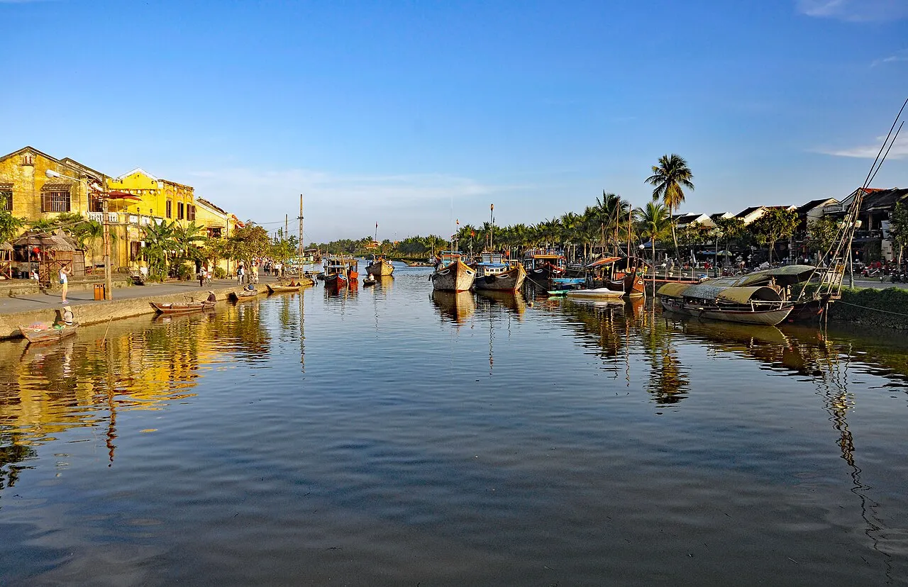Une rivière calme sous un ciel bleu, avec des bateaux amarrés le long des berges, des maisons jaunes à gauche et des palmiers au loin reflétés dans l'eau.