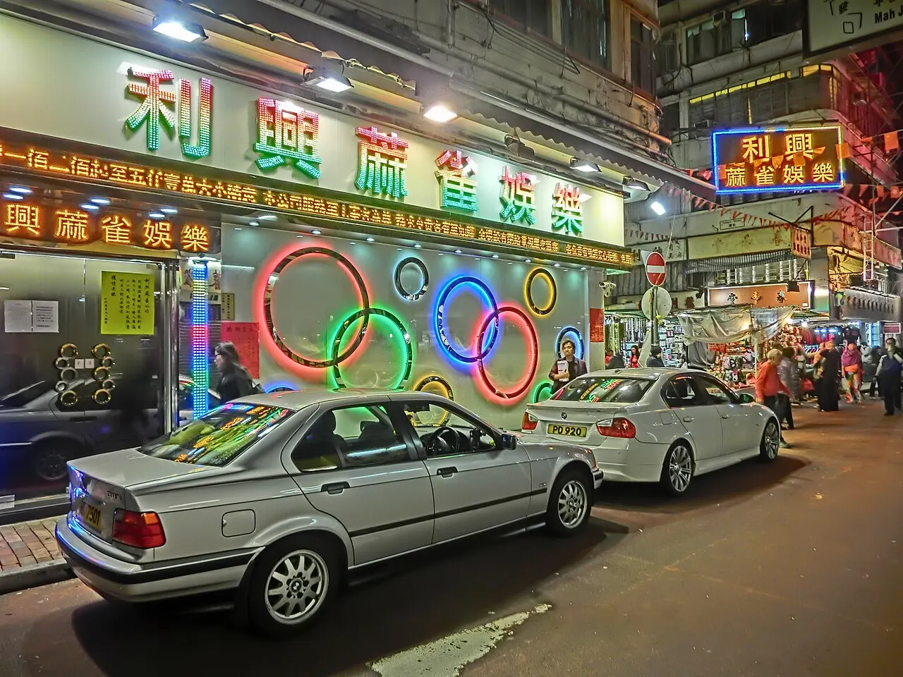 Rua da Temple Street à noite, com letreiros de neon, anéis luminosos e carros estacionados junto às lojas.