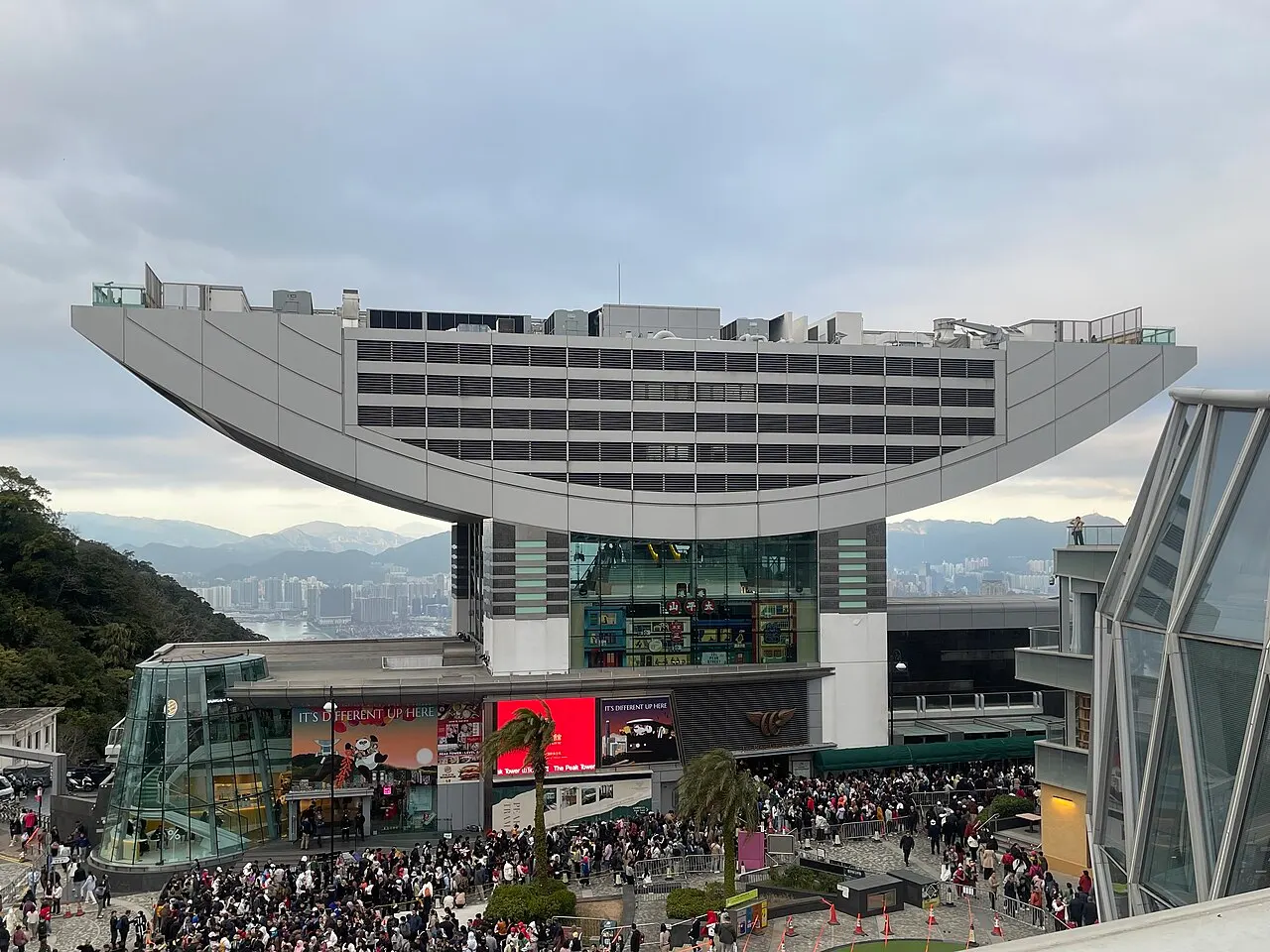 Edifício com cobertura curva em forma de tigela sobre um bloco de lojas, com gente na praça.