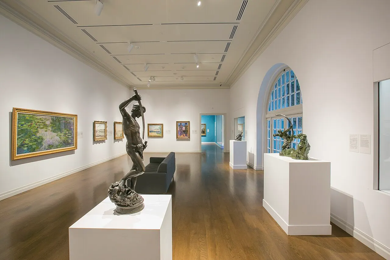 Uma galeria do Honolulu Museum of Art com piso de madeira, pinturas nas paredes brancas e uma escultura de bronze no centro.