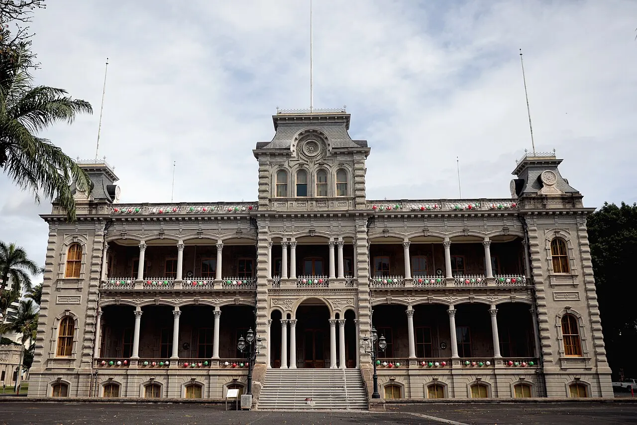 A fachada do ʻIolani Palace, com duas fileiras de varandas em arco, torres nos cantos e uma escadaria central, sob céu nublado.