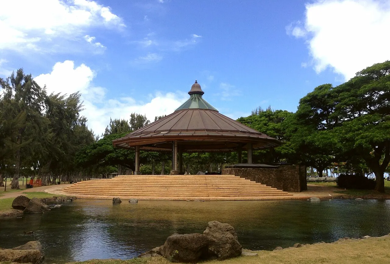 O coreto de telhado cônico do Kapiʻolani Park, à beira de um lago com pedras, entre árvores e sob céu azul.