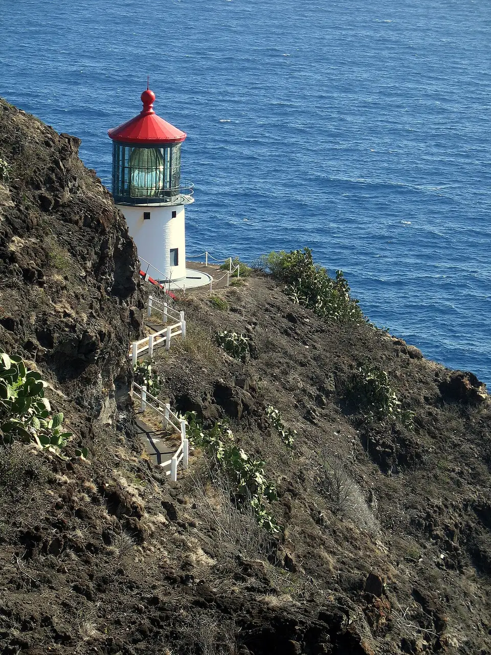 O pequeno farol branco de cúpula vermelha de Makapuʻu, encravado na falésia escura sobre o mar azul.