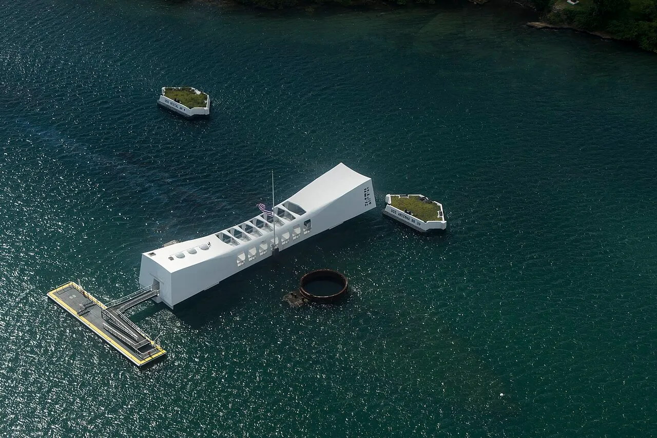 Vista aérea do memorial do USS Arizona, uma estrutura branca e longa sobre a água escura de Pearl Harbor, com o píer de embarque ao lado.