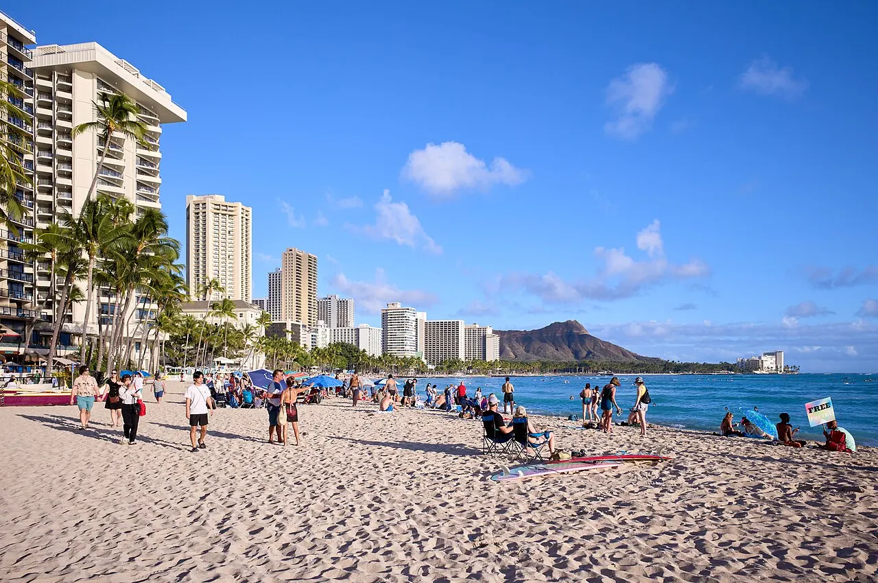A areia de Waikīkī cheia de banhistas, com hotéis altos e palmeiras à esquerda e o perfil do Diamond Head no horizonte.