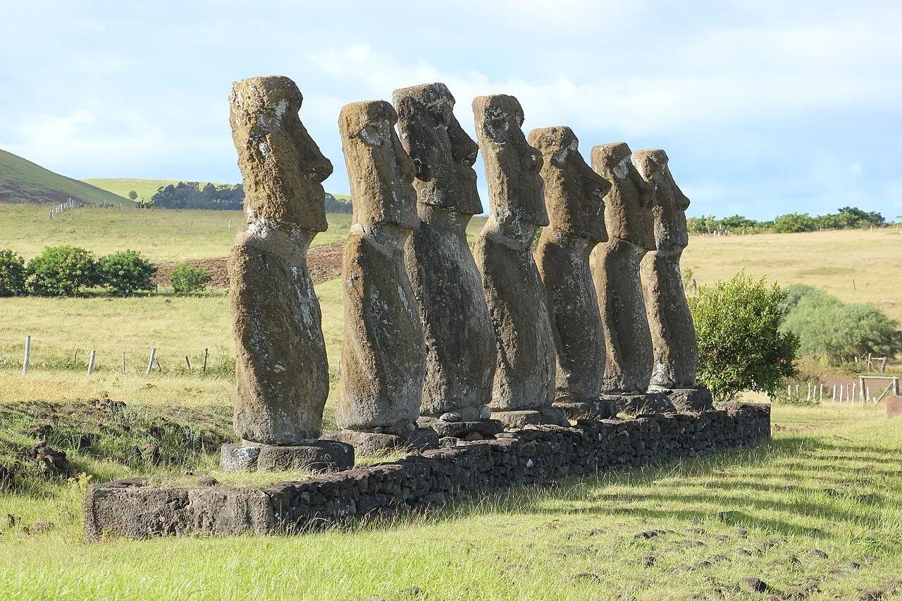 Os sete moais de Ahu Akivi enfileirados sobre a plataforma, vistos de lado num campo verde do interior da ilha.