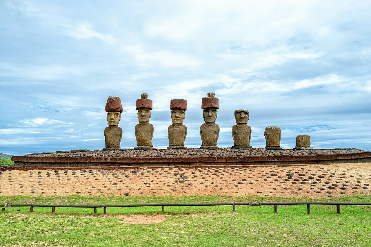 Sete moais sobre a plataforma de Ahu Nau Nau, quatro com o chapéu de pedra vermelha, diante de um gramado sob céu claro.