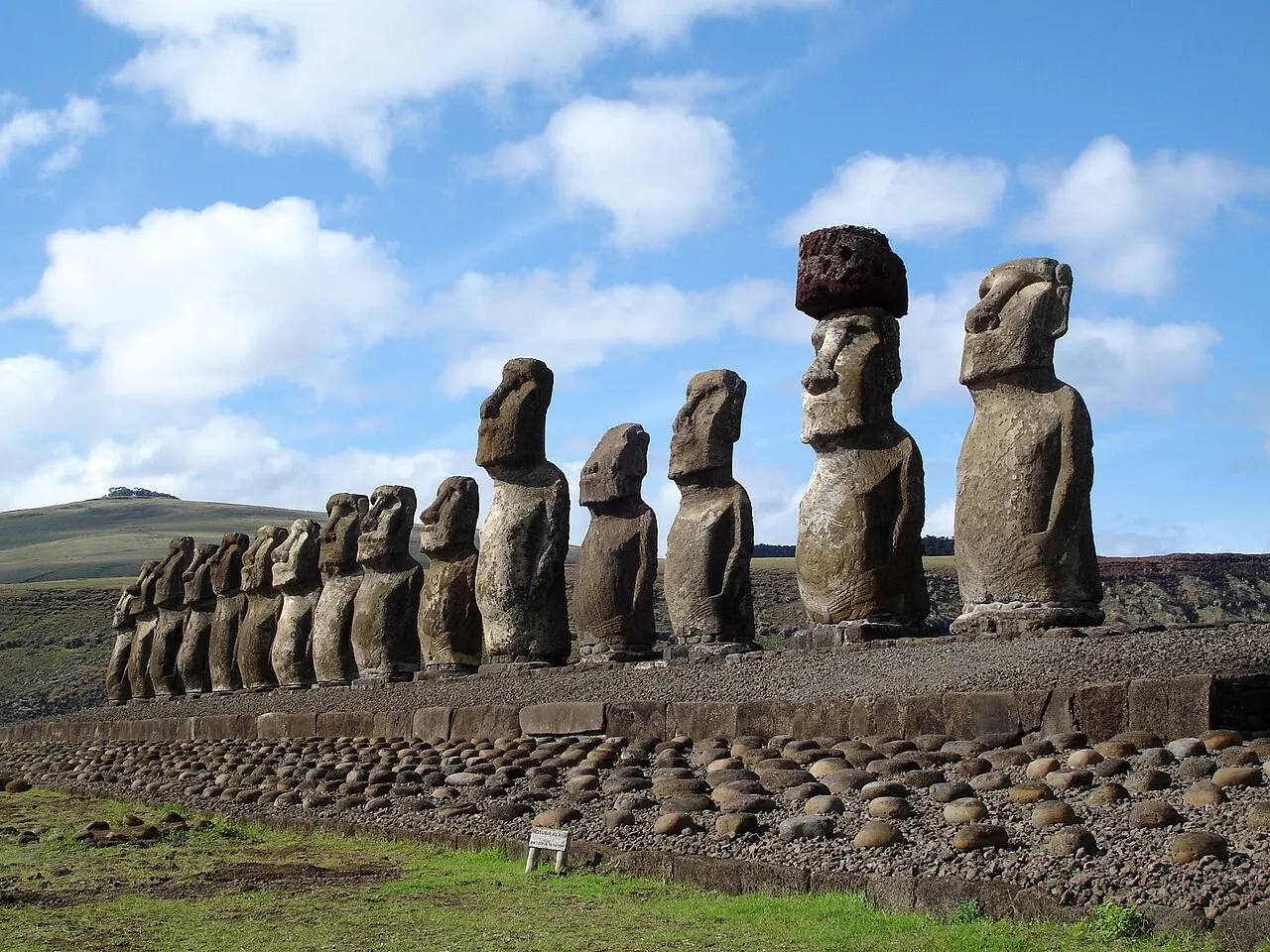 A longa fileira de moais de pé sobre a plataforma de Ahu Tongariki, um deles com o chapéu de pedra vermelha, e pedras redondas no chão à frente.