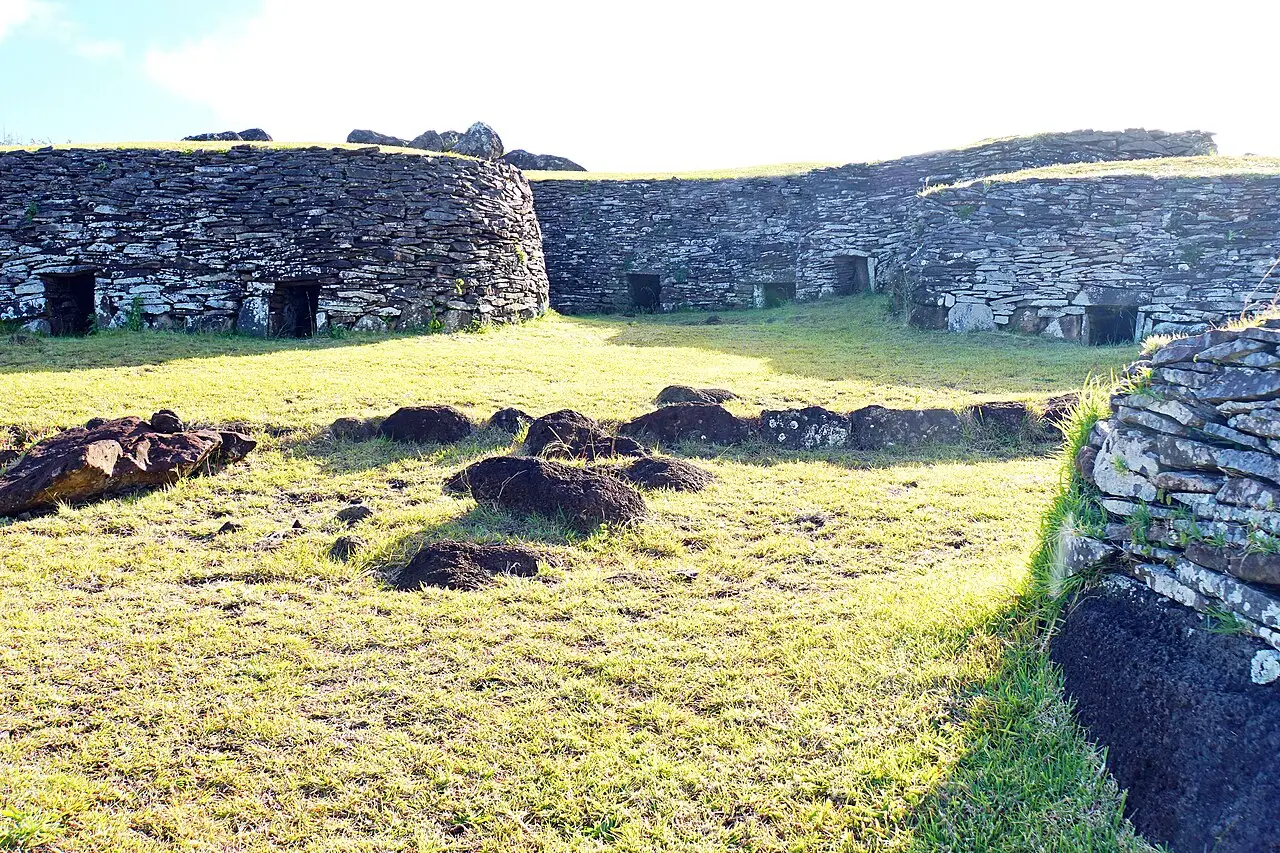 Casas baixas de lajes de pedra empilhadas, com entradas estreitas, em fila sobre o gramado da aldeia cerimonial de Orongo.