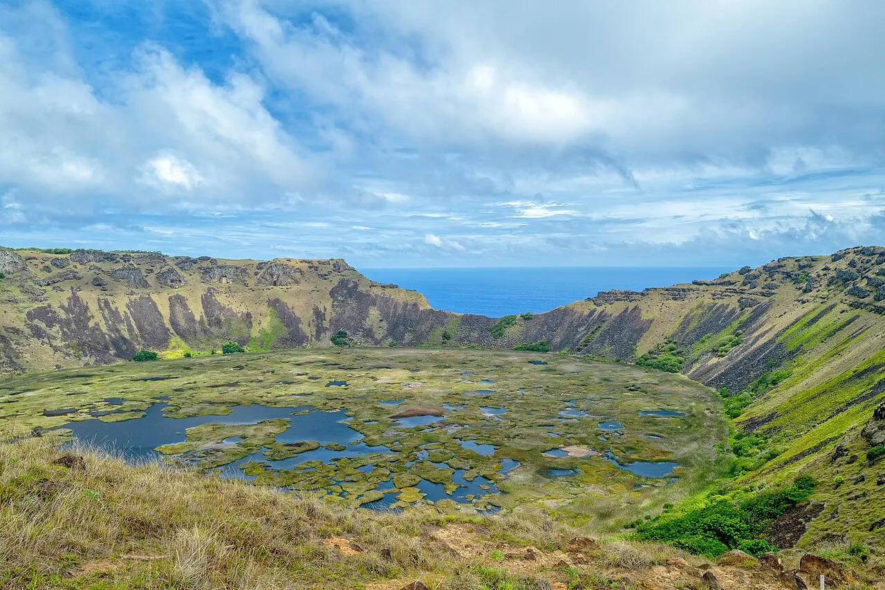 A cratera de Rano Kau vista da borda, com o fundo coberto de juncos e espelhos d'água e o oceano aparecendo por uma abertura da parede.