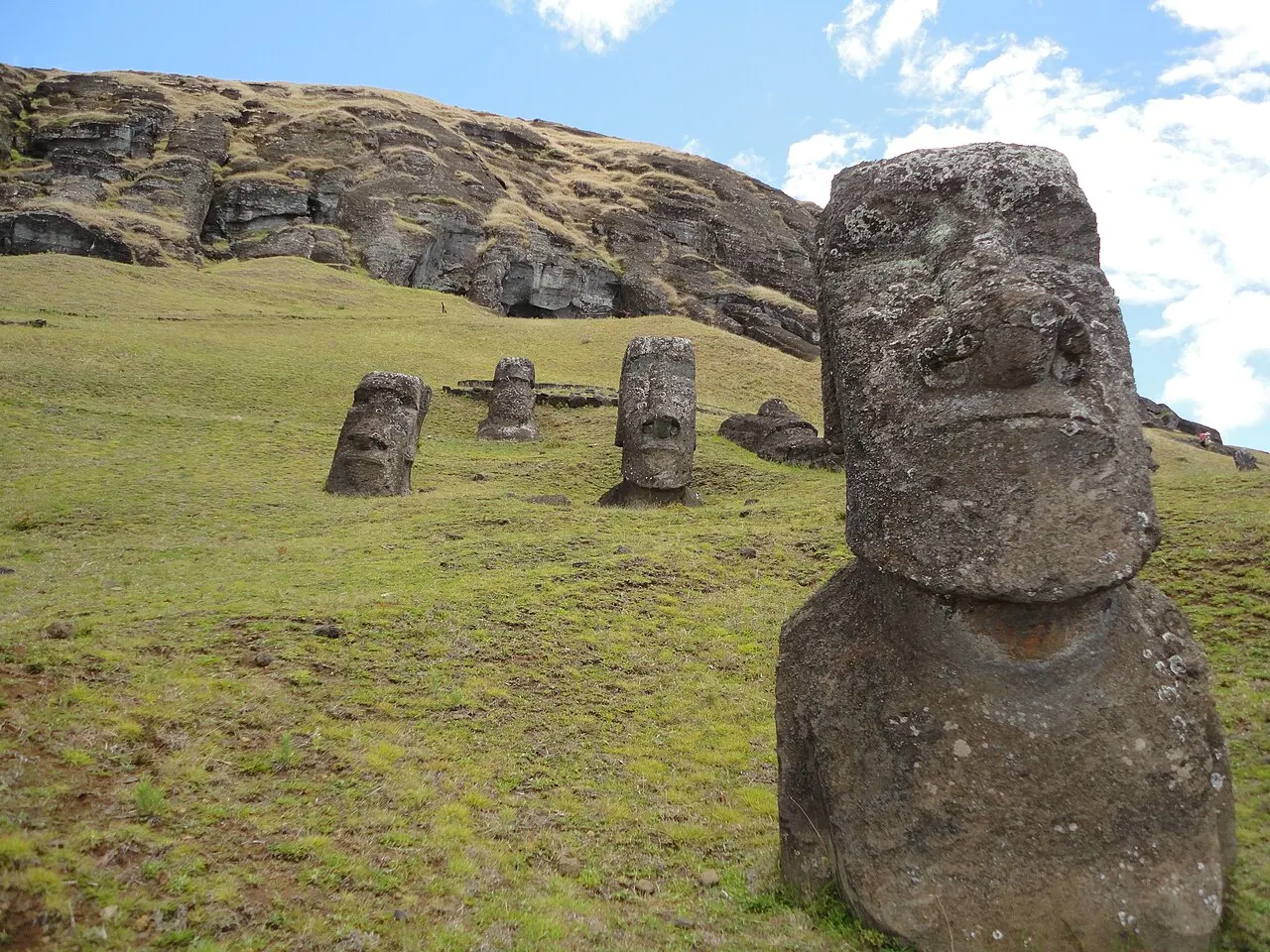 Cabeças de moai enterradas até o peito na encosta gramada de Rano Raraku, com a parede rochosa do vulcão ao fundo sob céu azul.