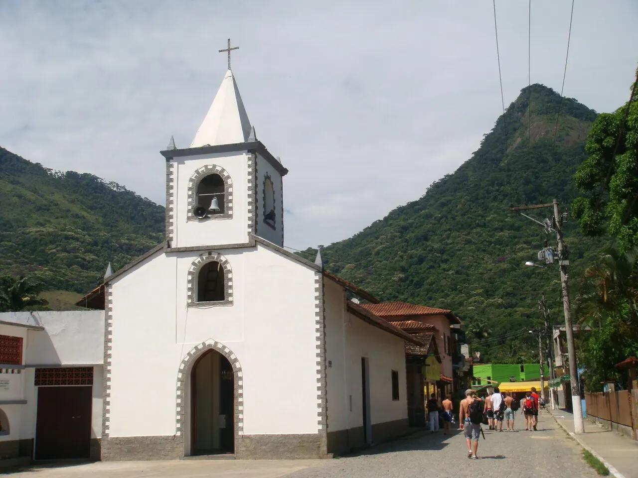 A fachada branca da Igreja de São Sebastião, com a torre do sino, na Vila do Abraão, com o morro verde atrás.