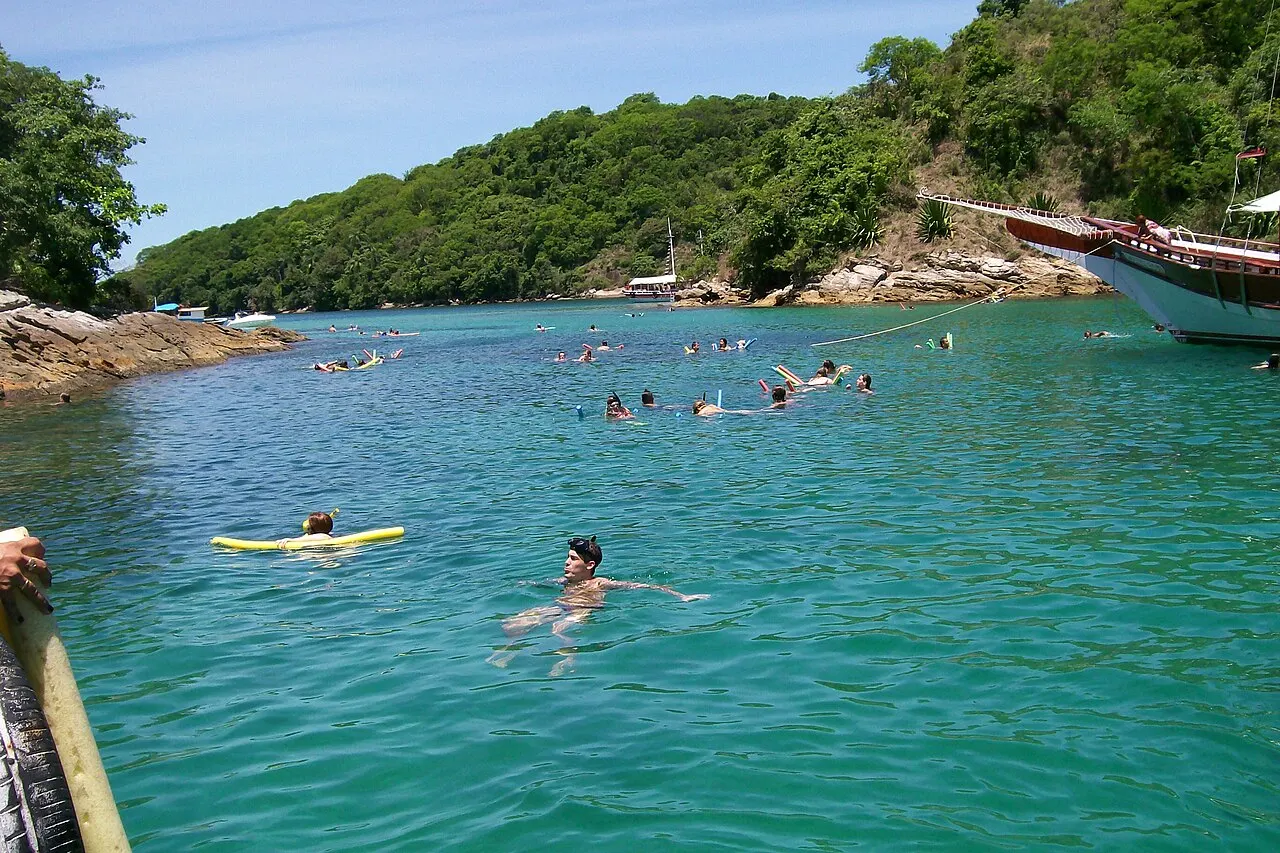 Banhistas nadando na água verde e transparente da Lagoa Azul, entre a ilhota e a costa coberta de mata.