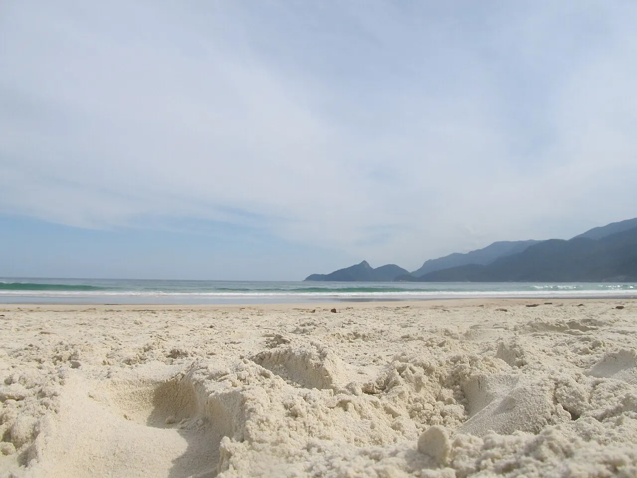 A areia branca e fofa da Praia de Lopes Mendes, com o mar e a linha de morros da ilha ao fundo, sob céu nublado.