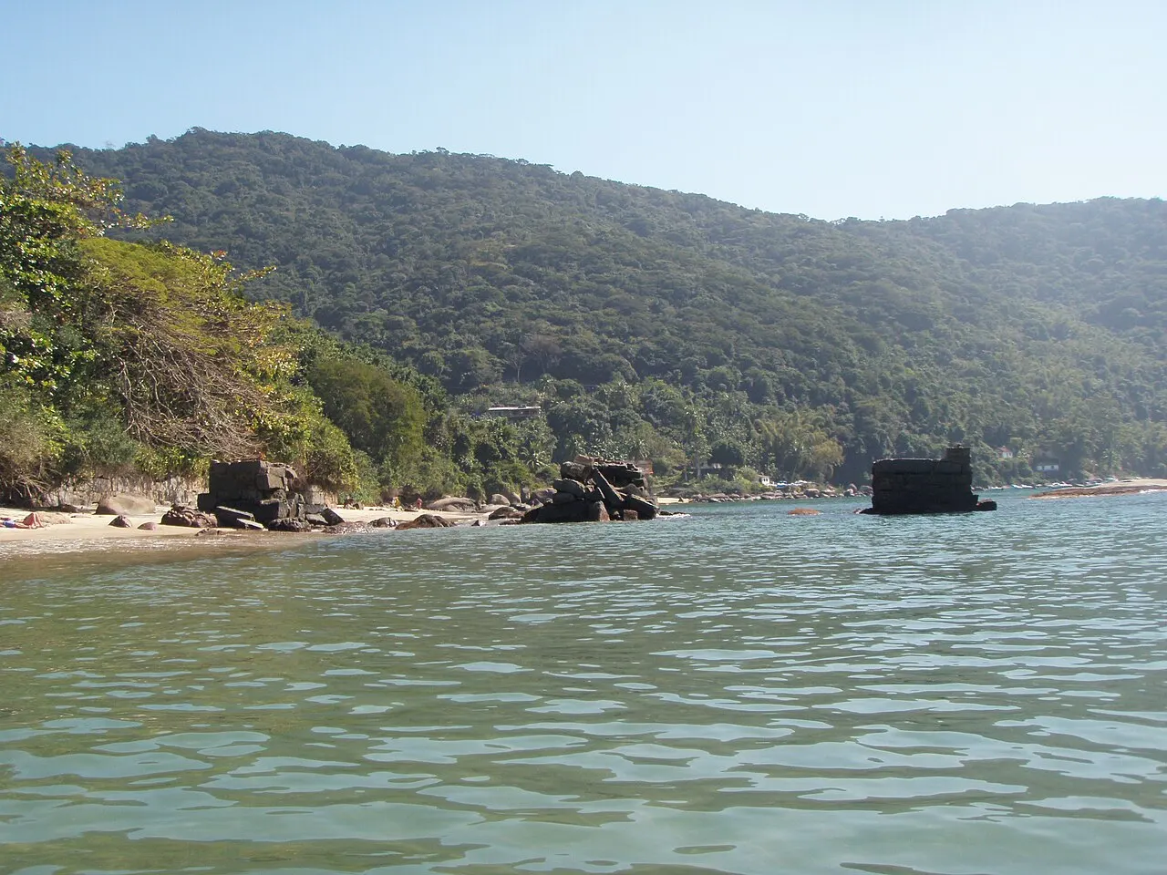 A enseada do antigo Lazareto com restos do cais de pedra na beira da água e o morro coberto de mata ao fundo.