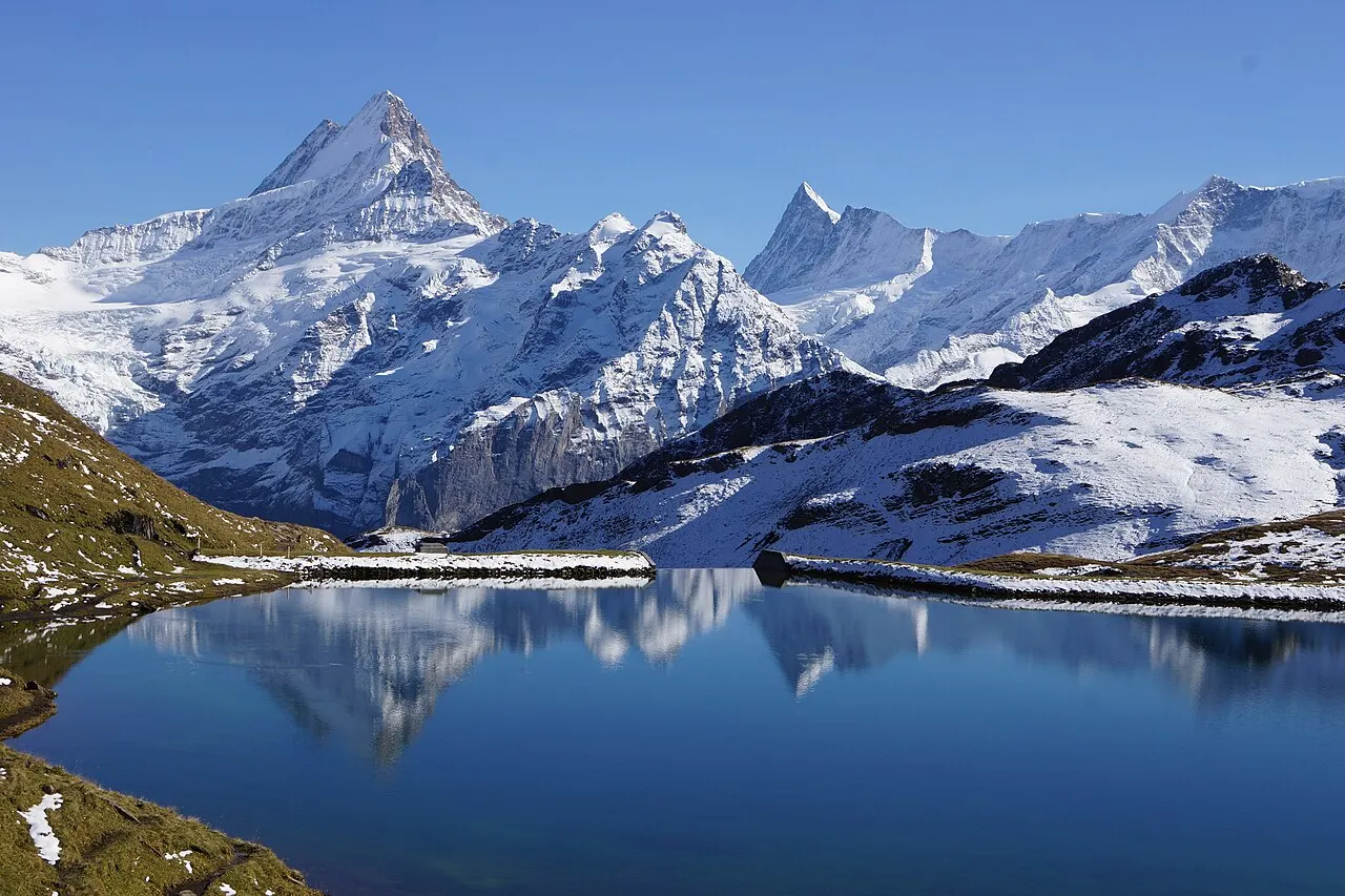 O lago Bachalpsee, de água azul e parada, refletindo os picos nevados do Schreckhorn e vizinhos, com neve nas margens.