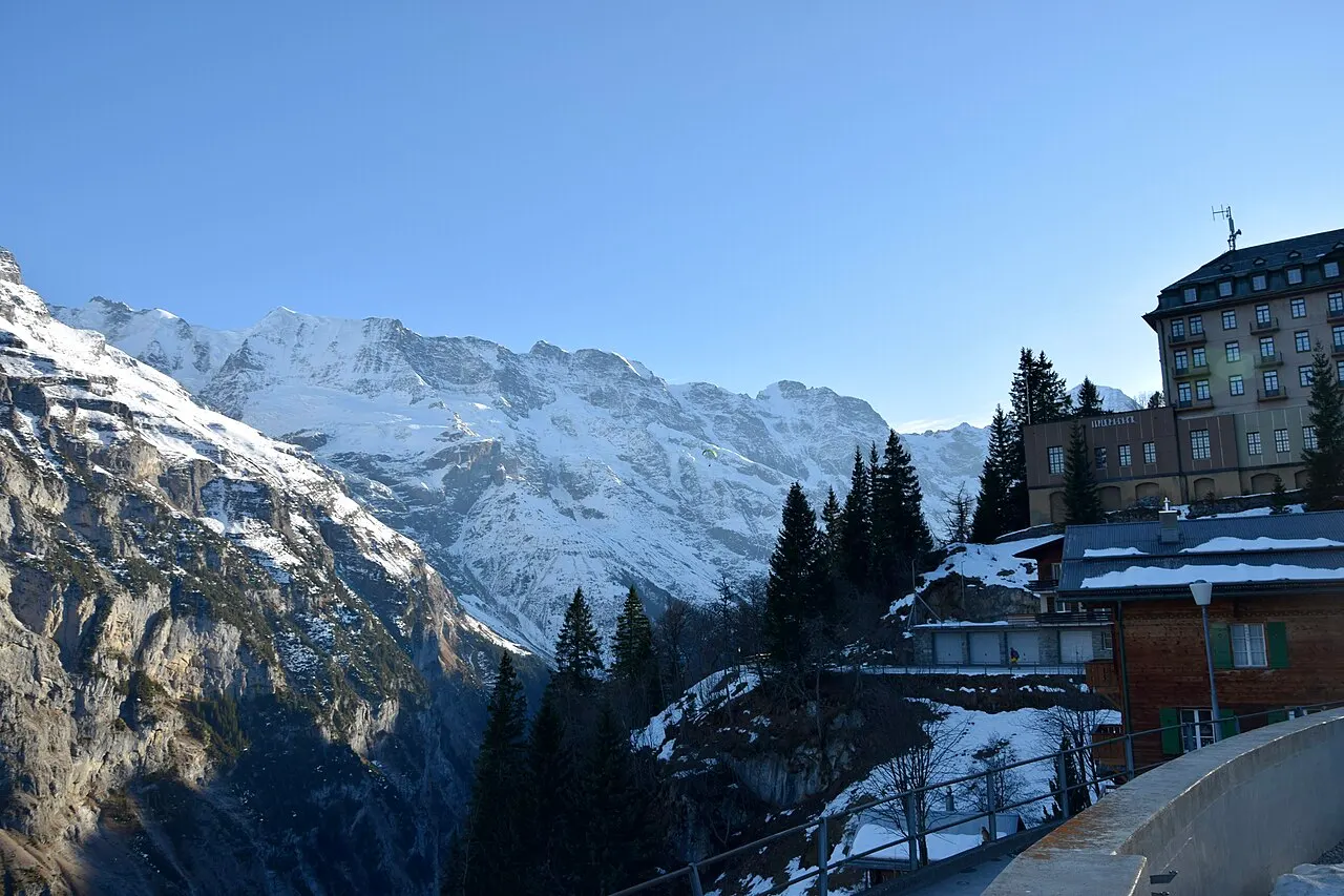 Os prédios de Mürren na beira do penhasco, entre pinheiros, diante da cadeia de montanhas nevadas do outro lado do vale.