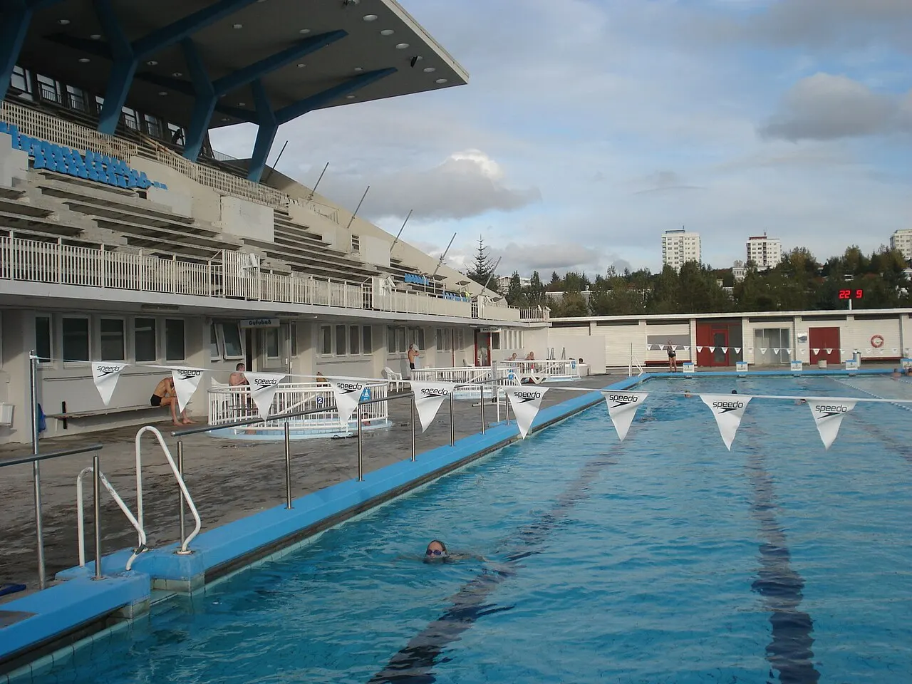 Uma piscina ao ar livre com raias demarcadas por boias, uma pessoa nadando, arquibancadas cobertas ao lado e prédios residenciais ao fundo, sob céu parcialmente nublado.