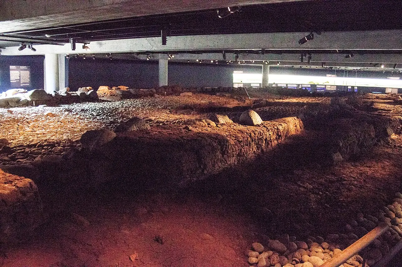 Os restos escavados de paredes de terra e pedra de uma antiga construção, iluminados por spots num ambiente escuro de museu, com pedras de rio dispostas ao lado e painéis informativos ao fundo.