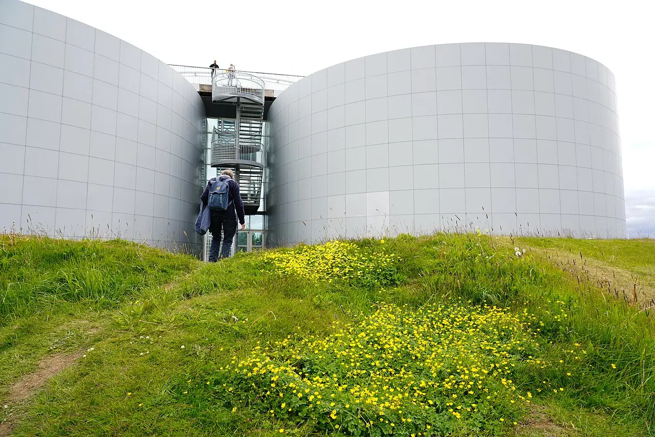 Dois reservatórios cilíndricos claros ligados por uma escada de metal, com flores amarelas e um visitante de costas no gramado.