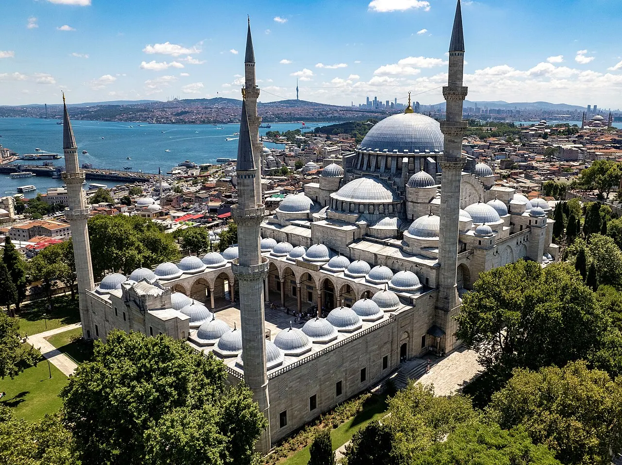 Vista aérea de uma grande mesquita com uma cúpula central cercada de meias-cúpulas menores e quatro minaretes altos e finos, pátio com arcadas e cúpulas pequenas em fileira, mar e cidade ao fundo, sob céu azul.