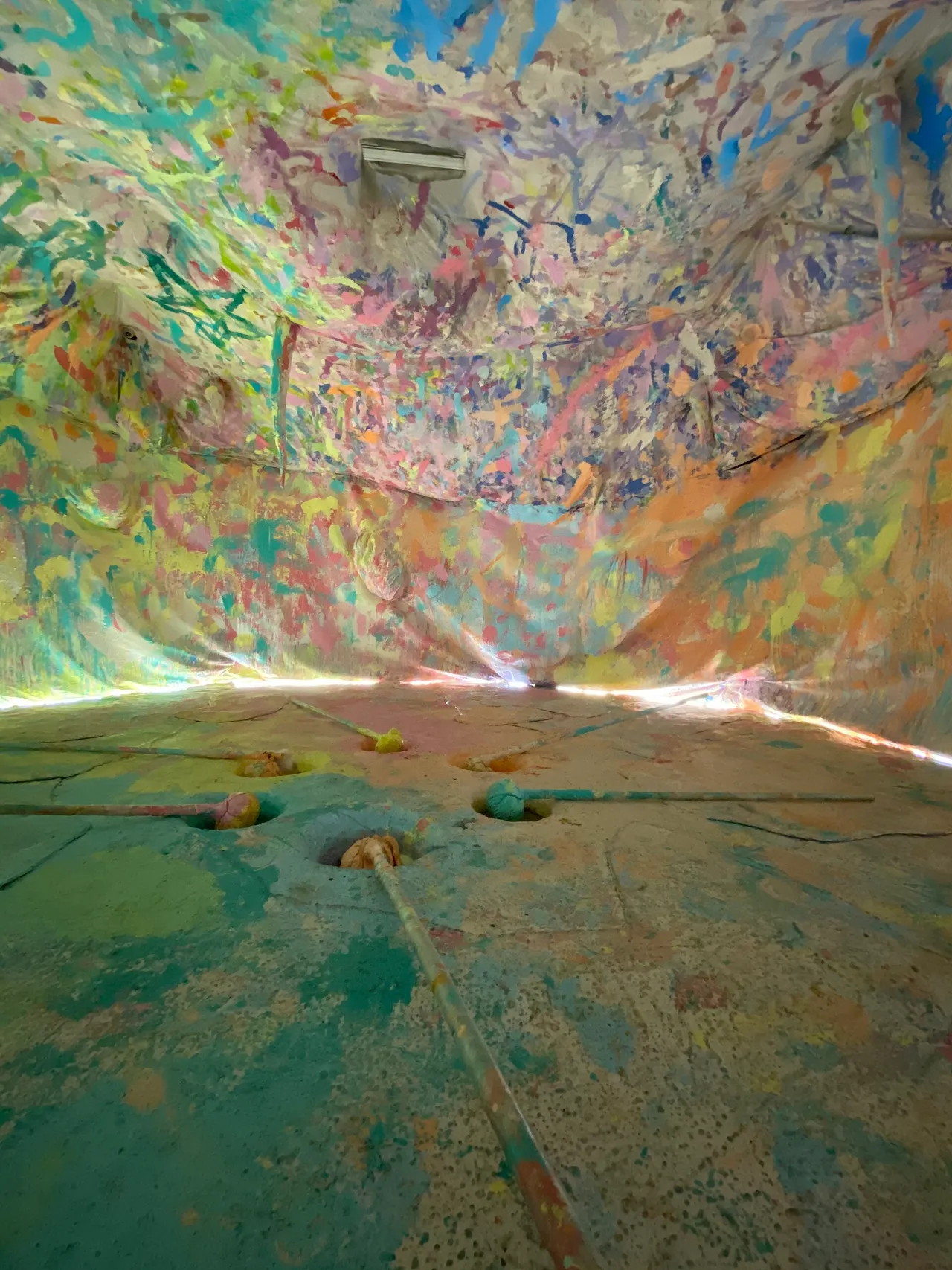Sala do Museum MACAN com teto, paredes e chão cobertos de tinta em muitas cores, e hastes saindo de furos no piso.