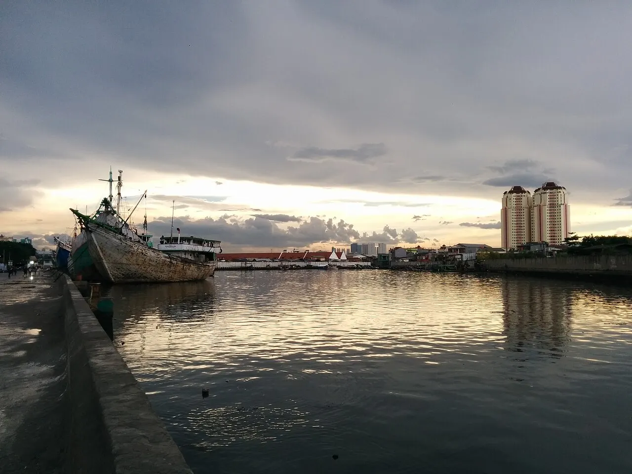 Bateau en bois au quai de Sunda Kelapa, avec l'eau du canal et de hauts immeubles au fond.