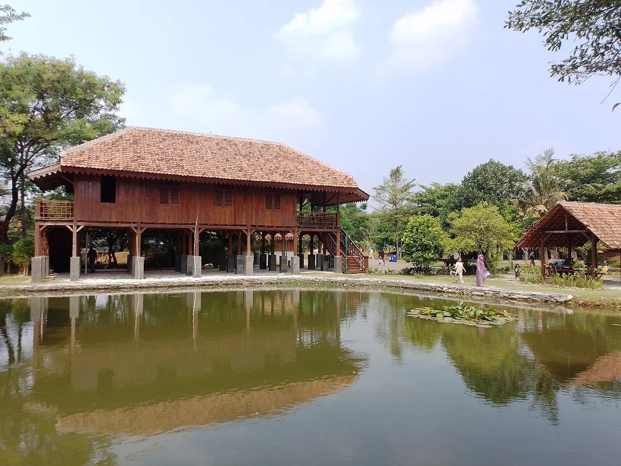 Casa de madeira sobre palafitas, telhado de telha, junto a um lago com árvores refletidas na água.