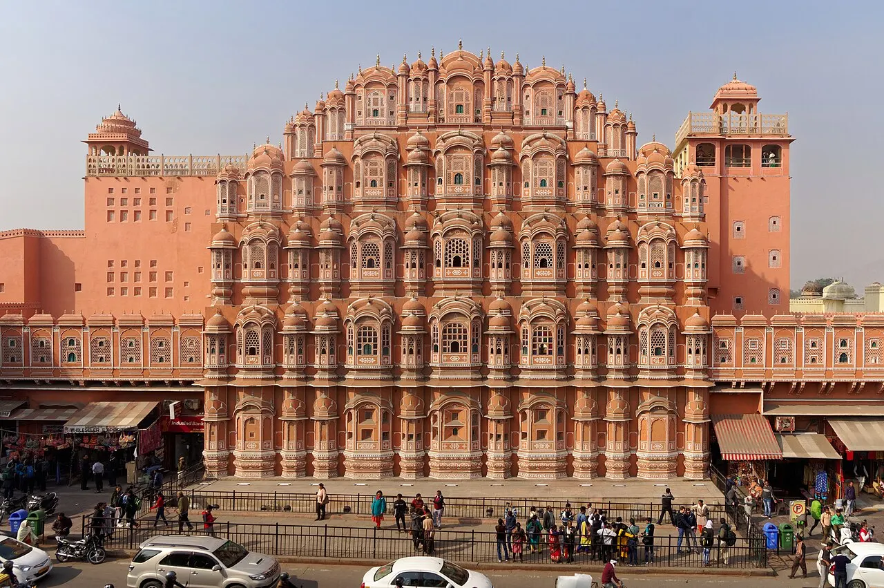 Fachada rosa inteira do Hawa Mahal, com as alas laterais e a rua com carros e pessoas na frente.