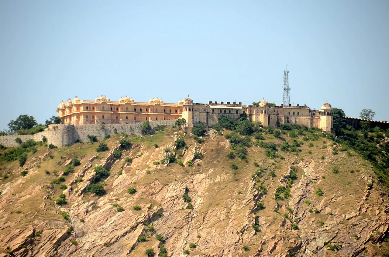 Um forte de pedra bege com ameias e pequenos domos decorativos, estendido ao longo do topo de uma colina rochosa e árida, com uma torre de transmissão metálica ao lado, sob céu azul claro.