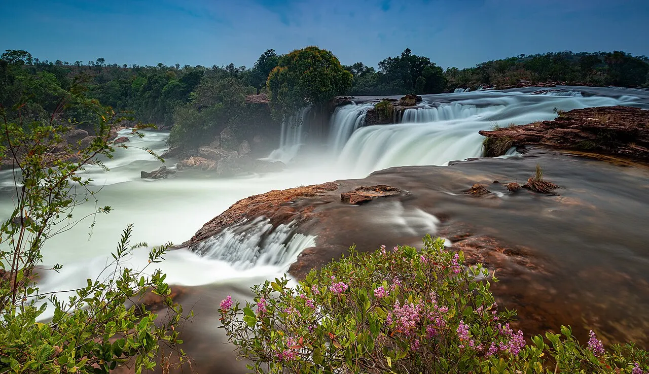 A Cachoeira da Velha, larga e em degraus de pedra, com flores rosadas na margem em primeiro plano.