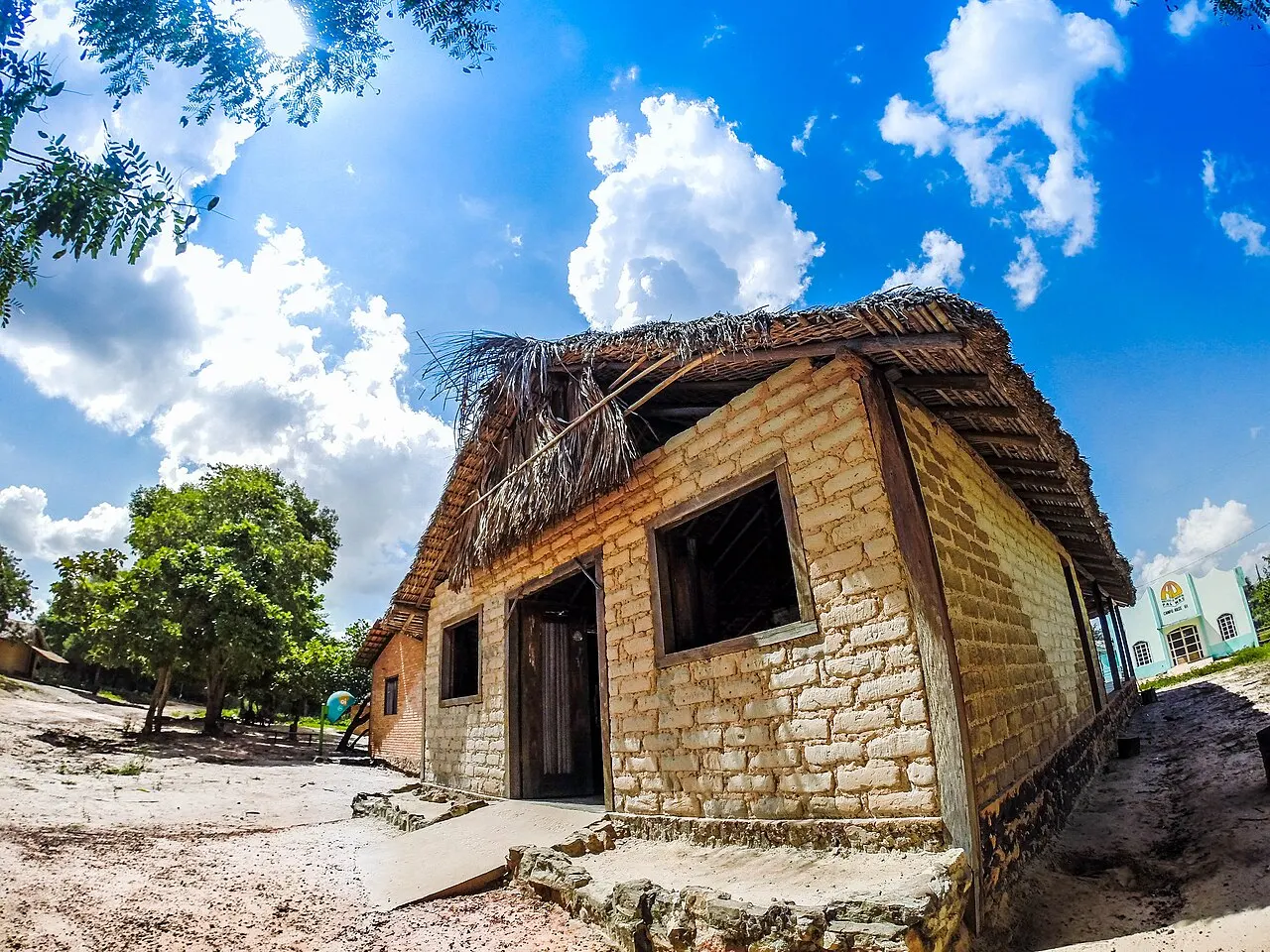 Uma casa de adobe com telhado de palha no povoado da Mumbuca, sob céu azul com nuvens.