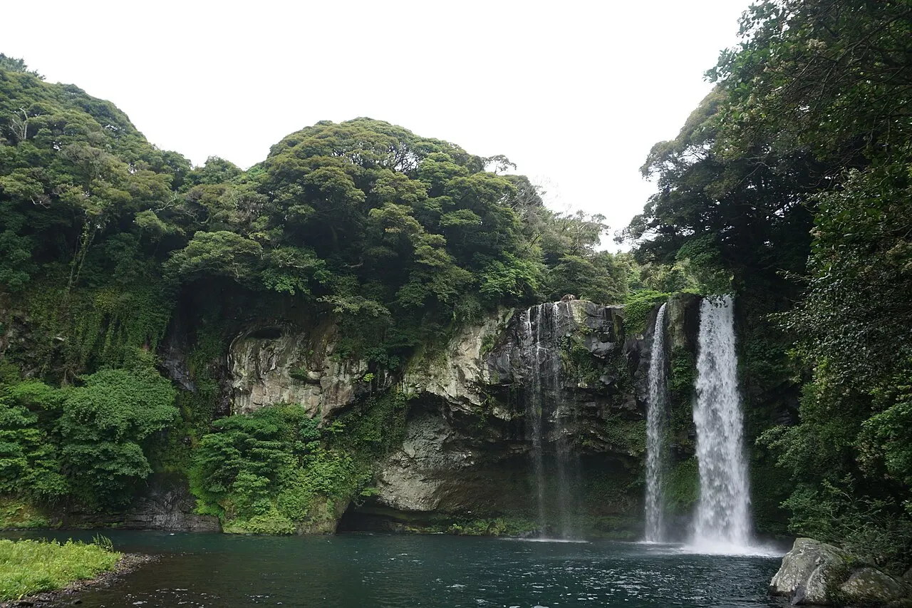La cascada de Cheonjiyeon cayendo en una poza de agua oscura, entre paredes de roca y árboles de follaje denso.