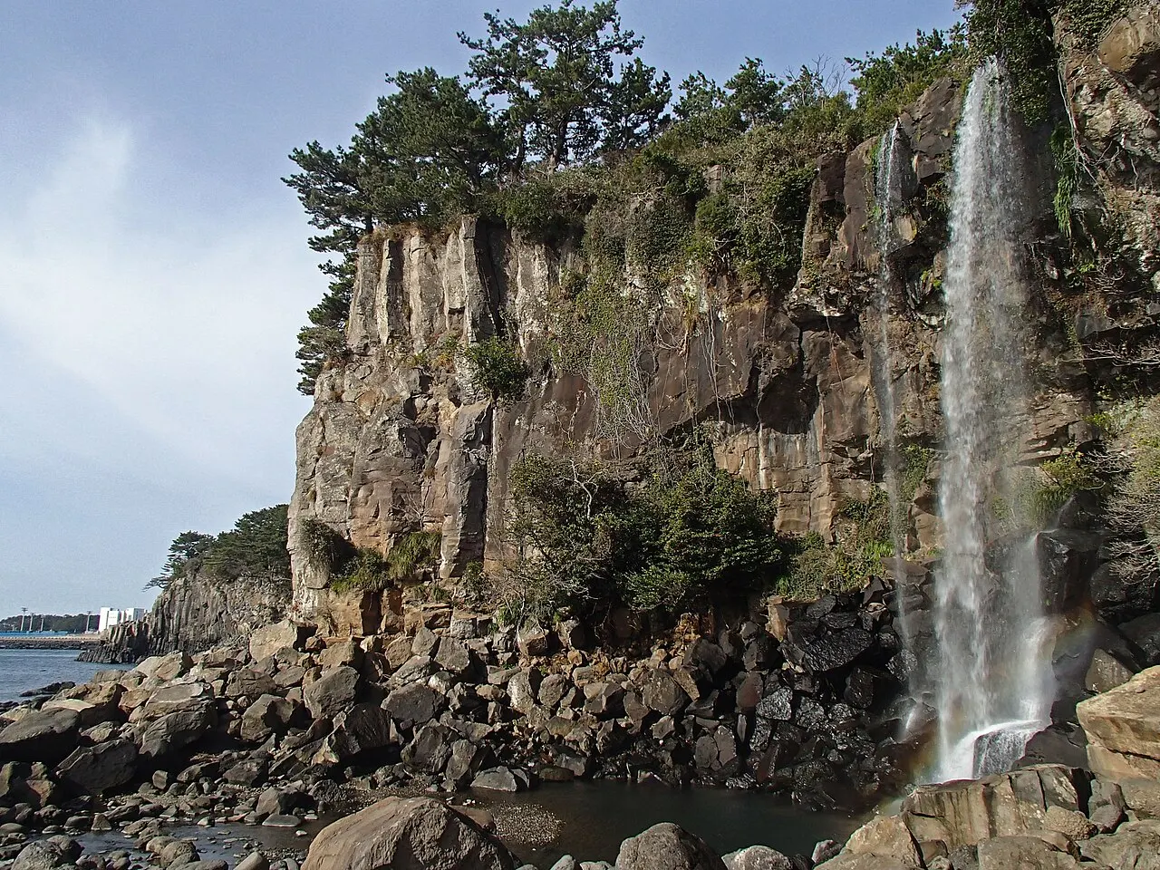 La cascada de Jeongbang cayendo de un acantilado de roca coronado de pinos sobre un lecho de piedras redondeadas junto al mar.