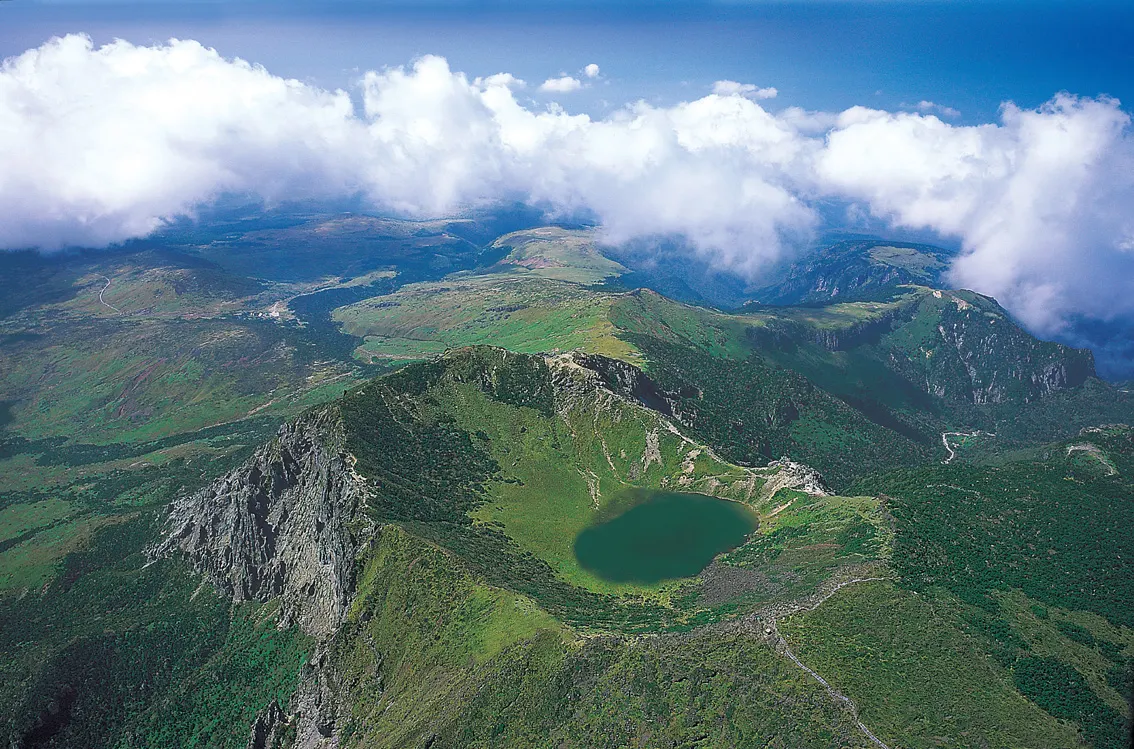 Vista aérea de la cima del Hallasan, con el lago verde en el fondo del cráter, laderas cubiertas de vegetación y nubes sobre el horizonte.