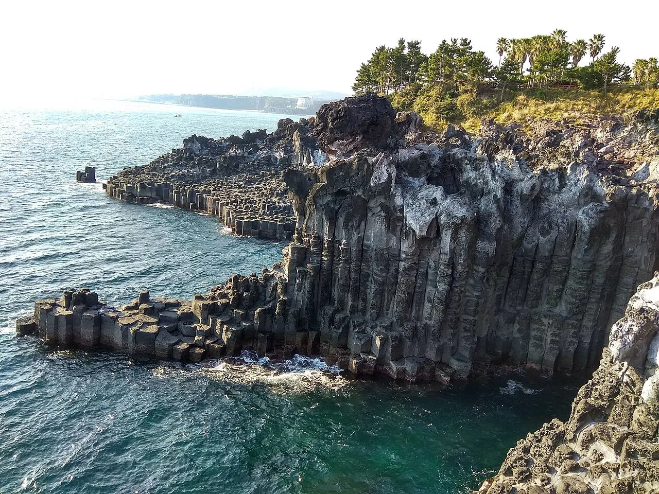 Pared de columnas de basalto hexagonales en Jusangjeolli, bajando hasta el agua verdeazulada, con árboles en lo alto del acantilado.