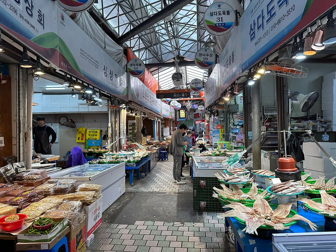 Pasillo cubierto del Mercado Dongmun, con puestos de pez sable y mariscos secos a la derecha, paquetes de alimentos a la izquierda y carteles colgados del techo.
