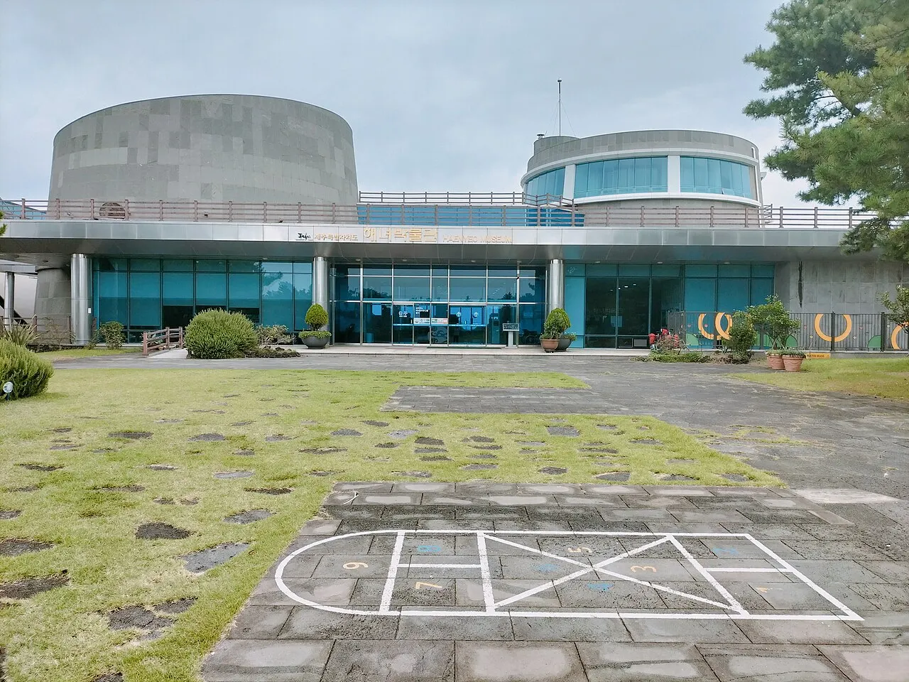 El edificio del Museo de las Haenyeo, de hormigón gris y vidrio azulado, con un césped y un patio de piedra delante.
