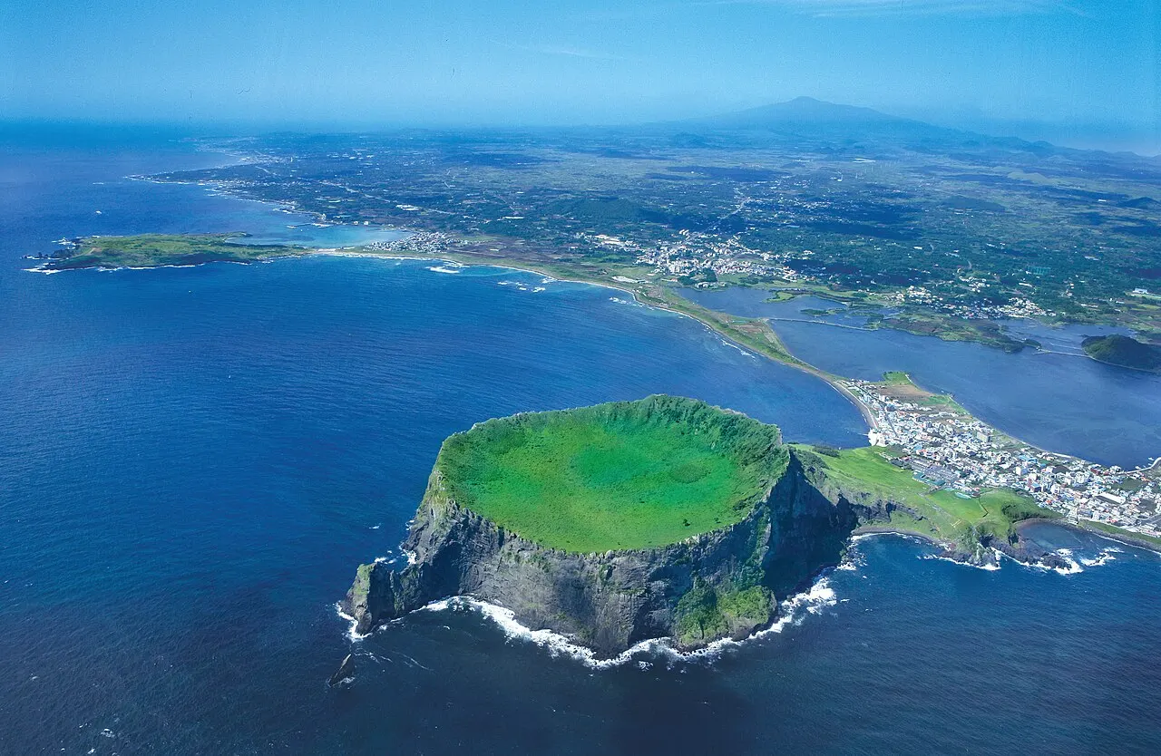 Vista aérea del cono de Seongsan Ilchulbong, con el cráter cubierto de hierba en lo alto, paredes oscuras sobre el mar y la franja de tierra que lo une a la isla.
