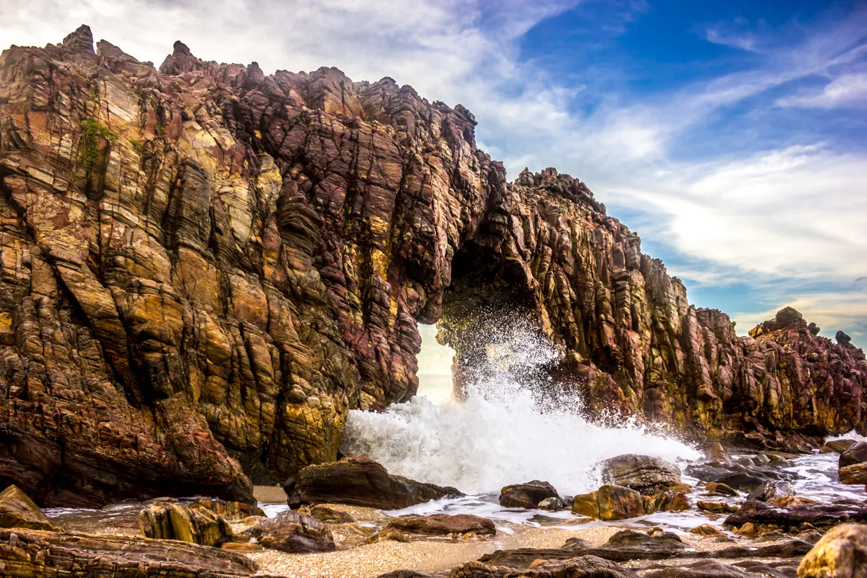 A Pedra Furada, em Jericoacoara, um arco de rocha escura e avermelhada na beira do mar, com uma onda arrebentando por dentro do buraco e céu azul com nuvens.