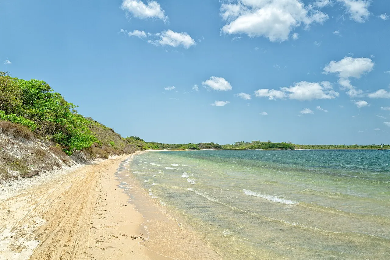 A Praia do Preá, com uma faixa de areia clara junto a uma encosta com vegetação, água rasa e transparente e céu azul com nuvens.