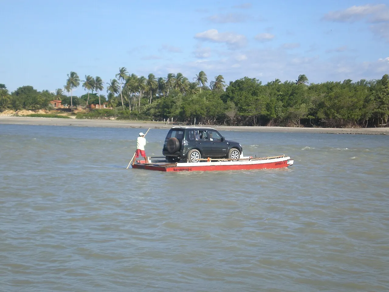 Uma balsa pequena levando um carro na travessia do rio Guriú, empurrada por um balseiro, com coqueiros e a margem verde ao fundo.