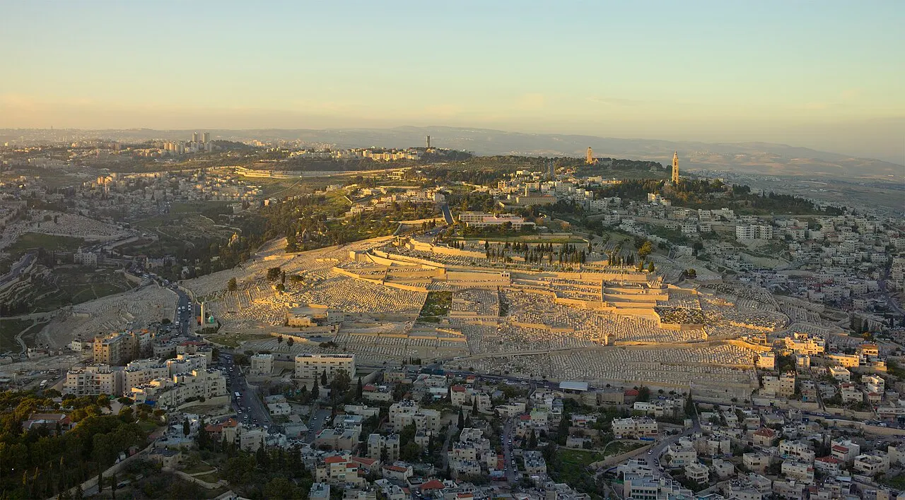 Vista aérea do Monte das Oliveiras na luz baixa do fim da tarde: fileiras de túmulos claros cobrem a encosta, com torres de igreja na crista e a cidade em volta.