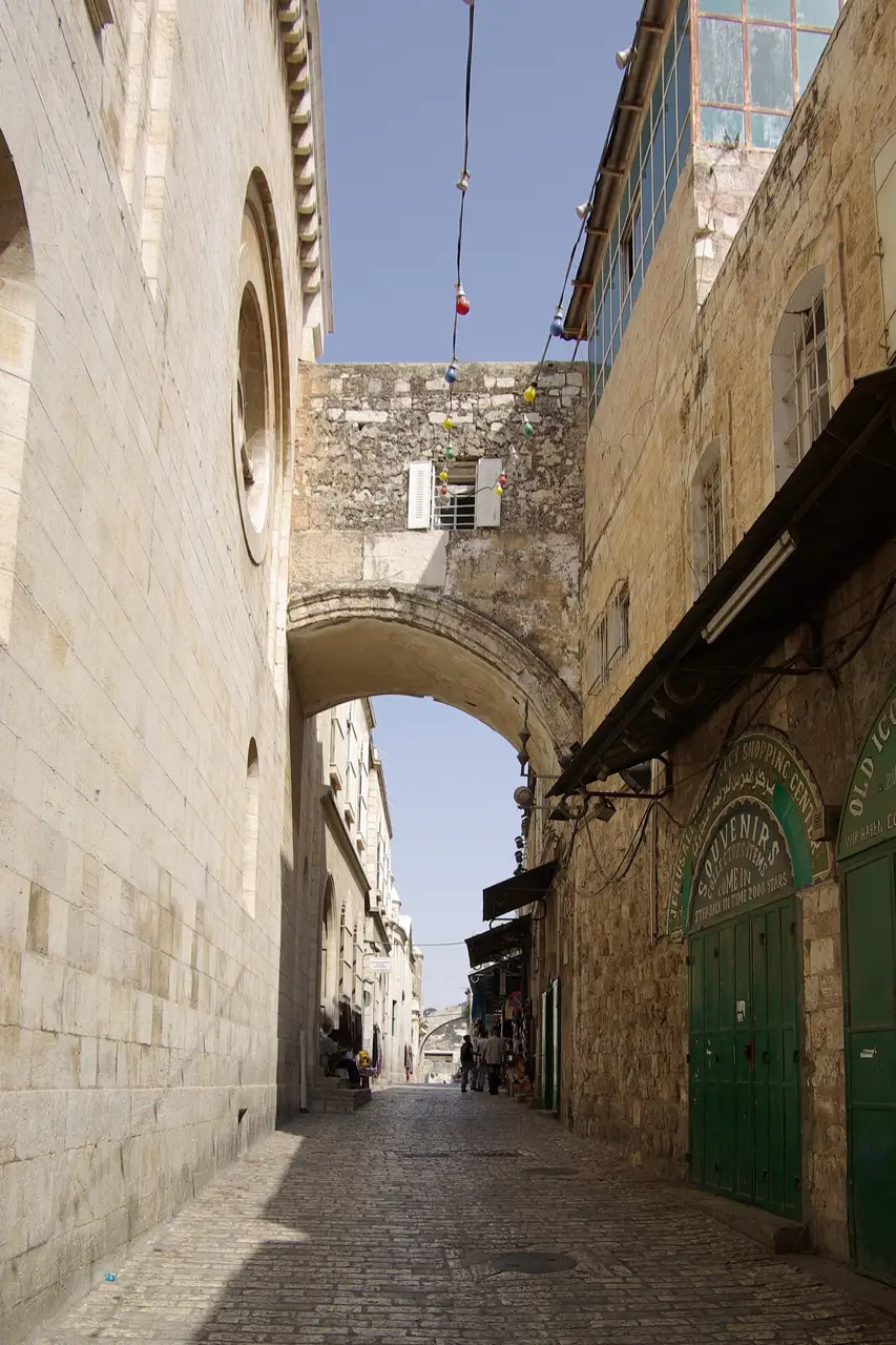 Arco de pedra da Via Dolorosa sobre a viela, com uma porta verde de loja à direita e poucas pessoas ao fundo.