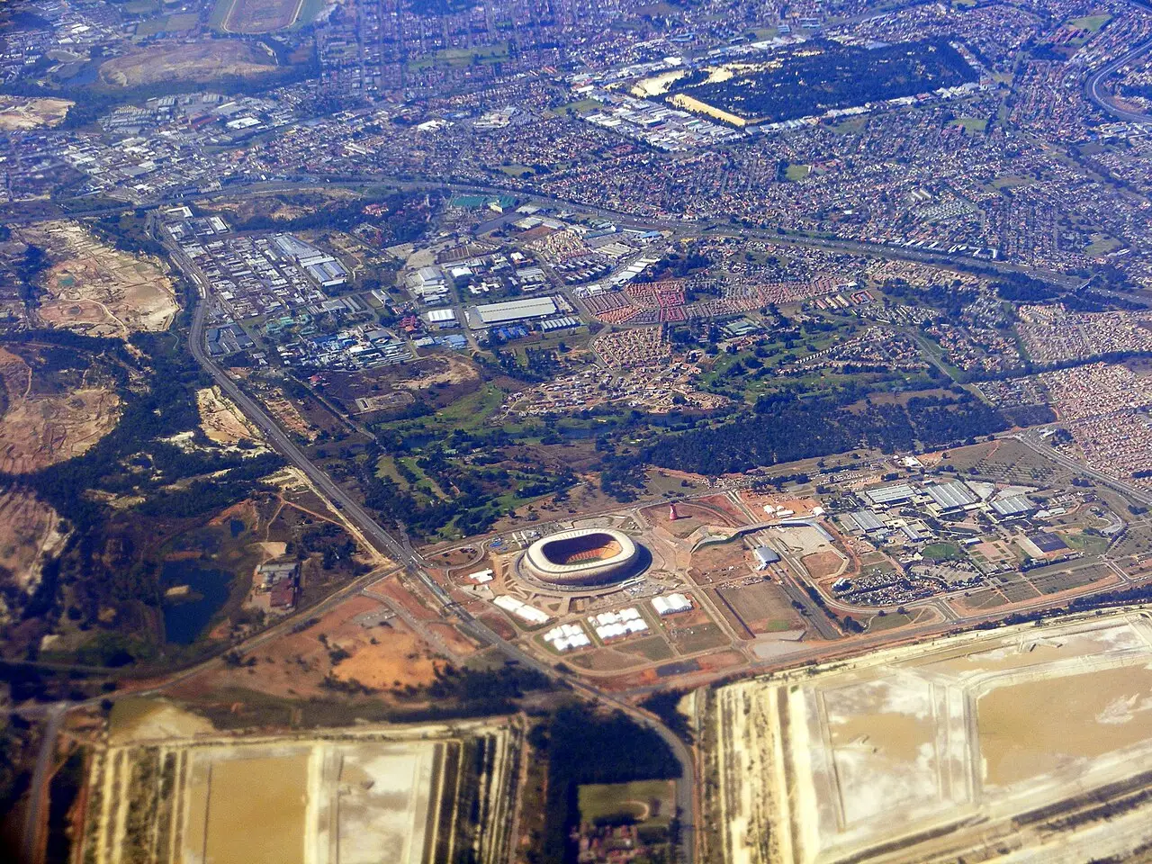 Vista aérea do Estádio FNB, oval e claro no centro do complexo, com a cidade e depósitos de minério em volta.