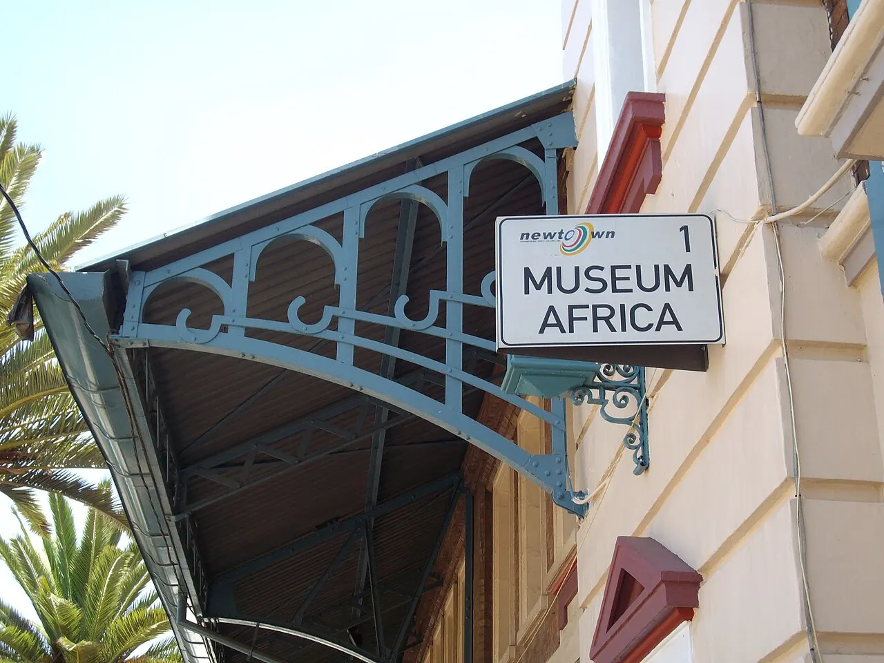 Placa branca com os dizeres “Museum Africa” presa a um suporte de ferro trabalhado na fachada clara do antigo mercado de Newtown, sob uma marquise de ferro azul e telha, com uma palmeira à esquerda.