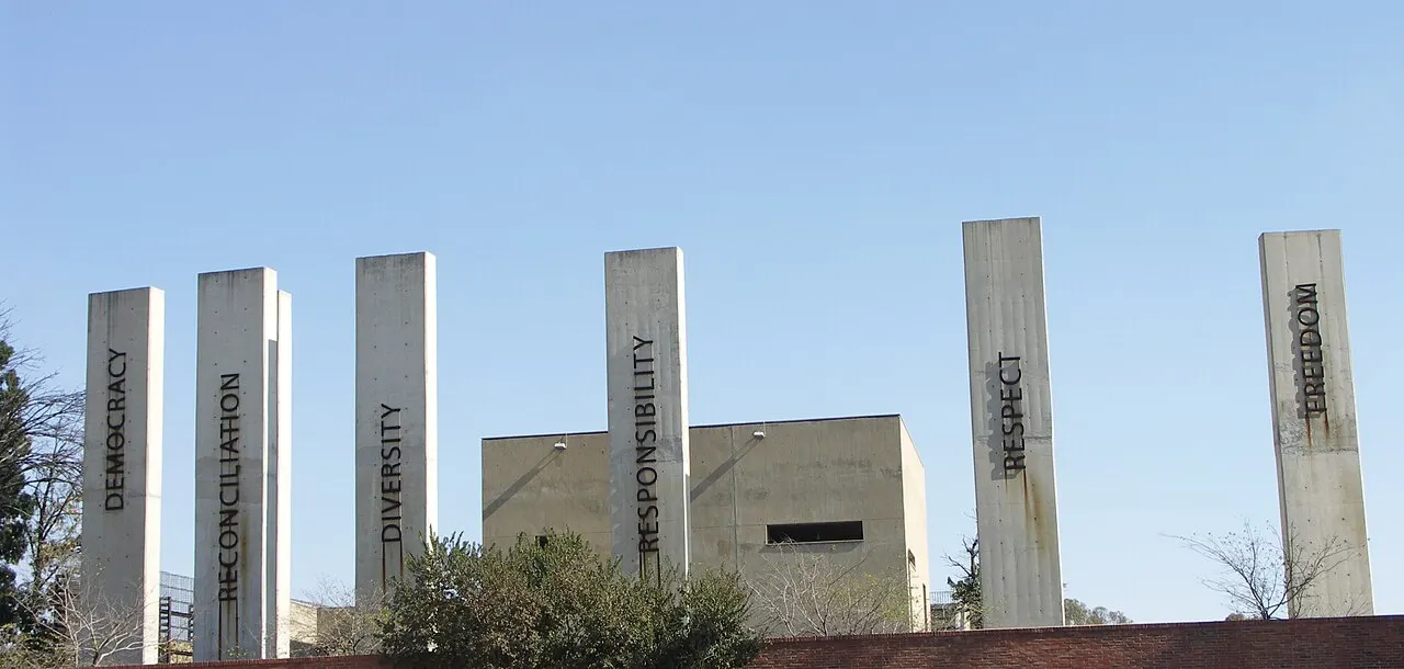 Sete altas colunas de concreto, cada uma gravada com uma palavra diferente em letras maiúsculas, junto a um prédio baixo de concreto e árvores, sob céu azul limpo.