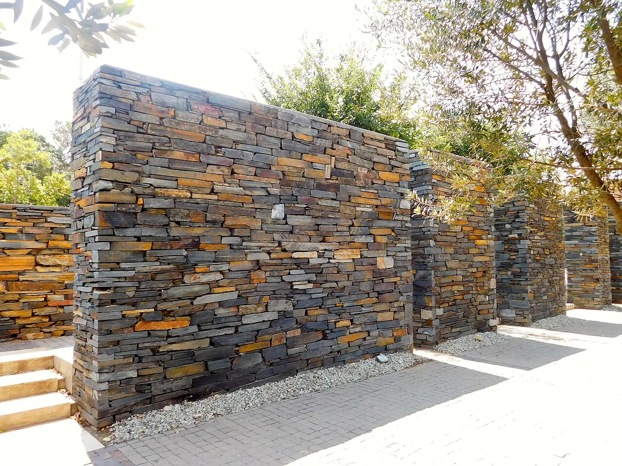 Um muro feito de pedras planas empilhadas em tons de laranja, cinza e marrom, formando painéis separados por pilares, com degraus de concreto e árvores ao lado, sob céu claro.