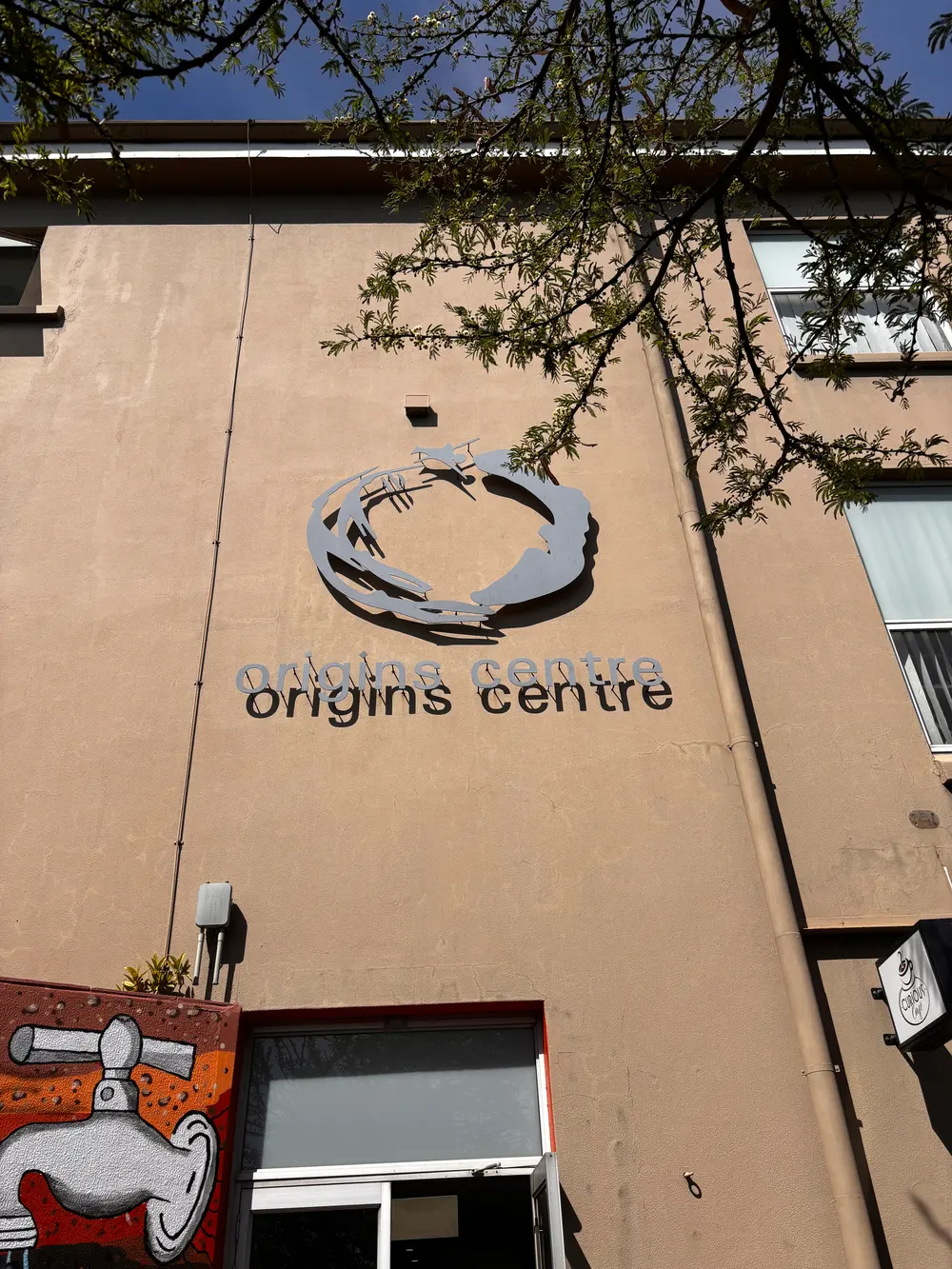 Fachada do Origins Centre com o nome do museu em letras metálicas, símbolo, janelas e ramos de árvore em primeiro plano.