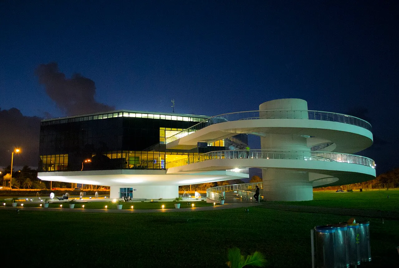 O prédio da Estação Cabo Branco iluminado à noite, com a rampa em espiral branca e o gramado na frente.